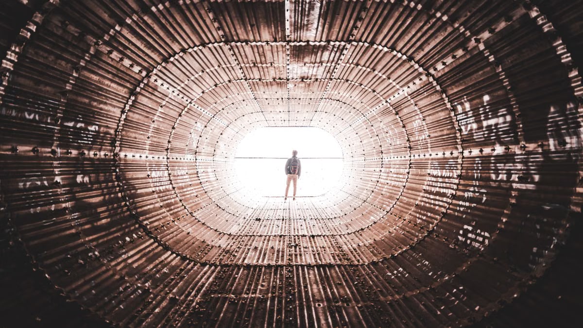 Silhouette of a person standing at the end of a geometric, metallic tunnel with dramatic perspective and lighting.